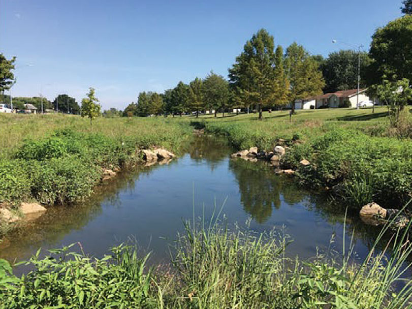 A subwatershed creek surrounded by rocks and vegetation. Homes are visible in the distance.