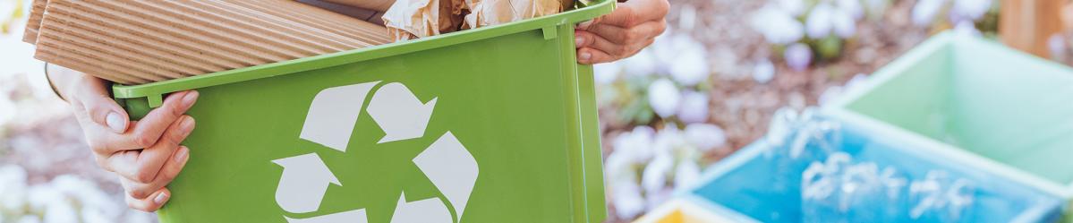 A woman holding a green tub with the recycling logo, standing next to three recycling bins for separating recyclables