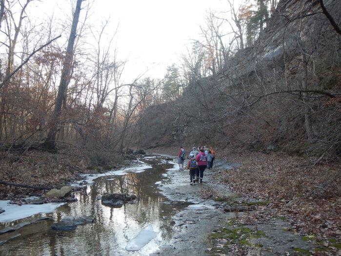 Group of adults and children hiking along a stream and cliff.