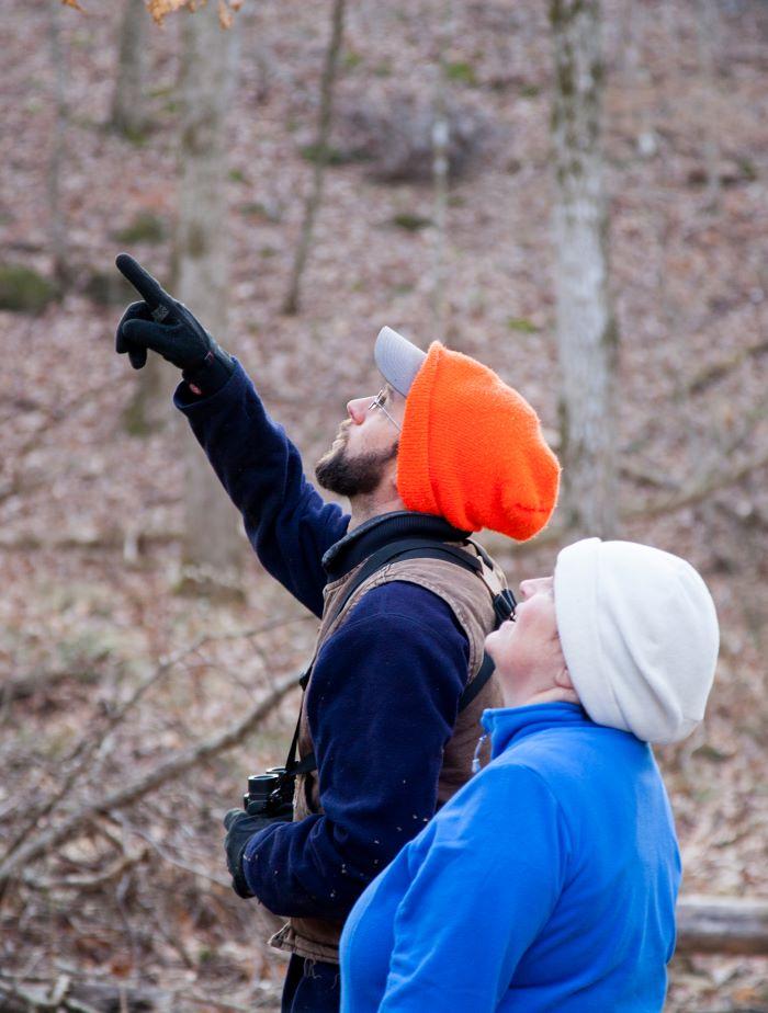 Man in jacket, coveralls and stocking hat pointing to a bird while holding binoculars. A woman in coat and stocking hat looks on.