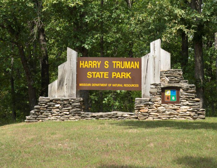 Brown cantilever sign that says “Harry S Truman State Park” with stone on both sides in front of trees.