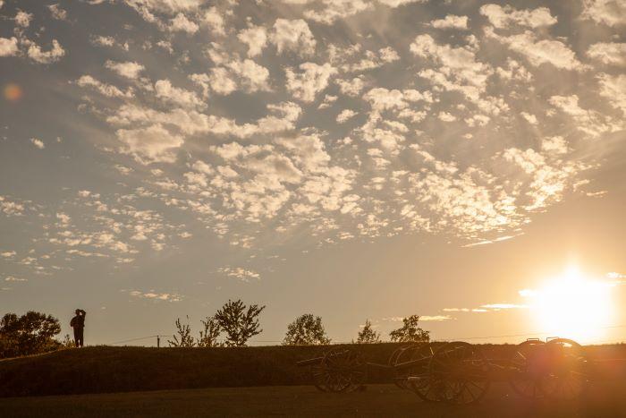 Sun setting over a hill; man saluting with 2 cannons in foreground.