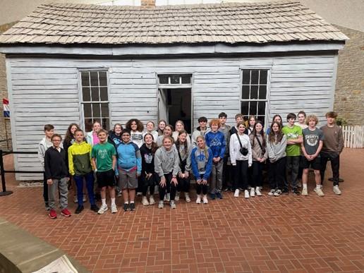 Group of 30 young students stand in front of old cabin that has 2 windows and an open door.