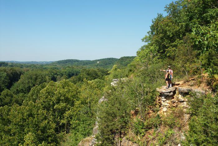 Two people standing on a bluff, wearing shorts, t-shirts and hats are looking out over the landscape and one is pointing in the valley.