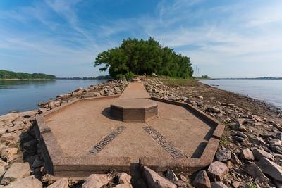 Concrete marker with a sidewalk leading to it with a river on either side.