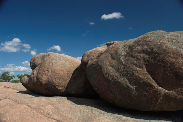 Two large rocks with trees in the background and blue sky with little white clouds.