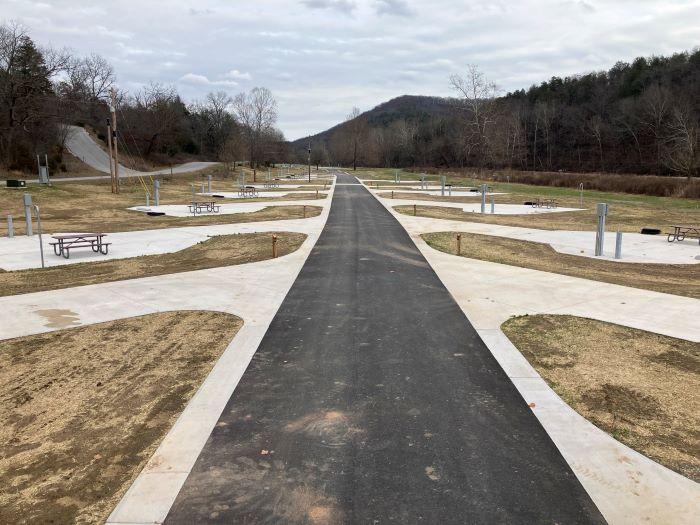 Asphalt road with campsites on the sides with picnic tables. Hills and trees in the background.