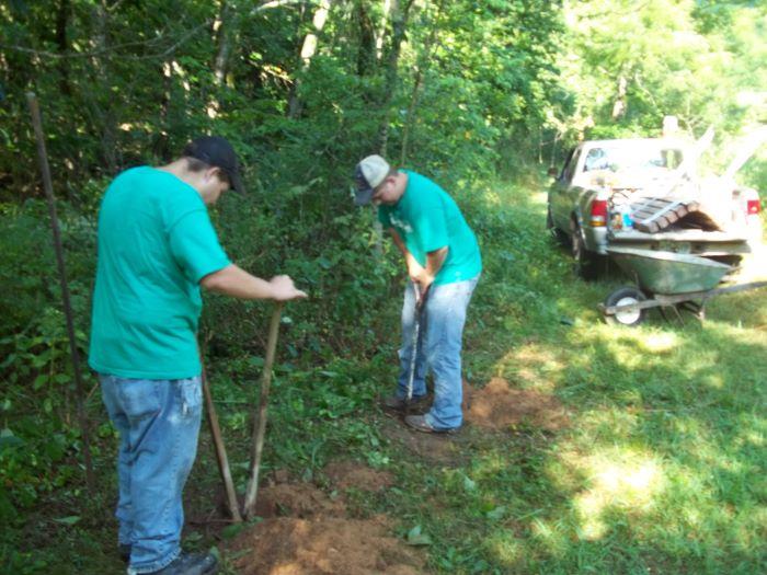 Two males in jeans and  t-shirts use tools to level the dirt on a trail. A green wheelbarrow and a pick-up truck are in the background.