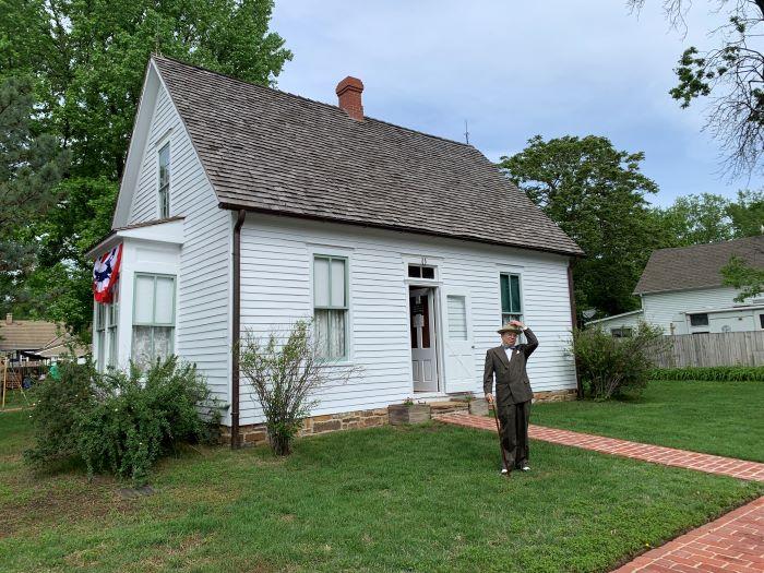 Man dressed in suit with his hand on his hat, standing outside of a small, two-story while house with a brick sidewalk leading to the door.