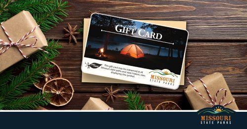 A Missouri State Parks gift card with a camping scene is displayed in a holiday setting with wrapped gifts and greenery.