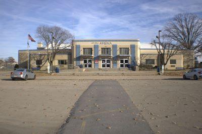Front view of the beige and blue arena building in cape Girardeau with trees, parked cars, and an American flag on a clear day 