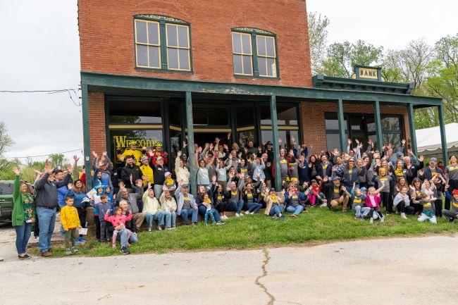 Group of people, children and adults, pose with raised hands in front of the Treloar Depot, a brick building with a 