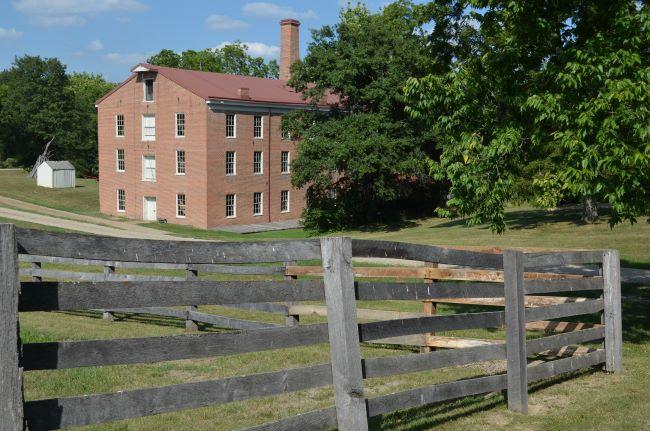 3 story brick building, red roof and chimney and small white shed nearby. Trees line the back and side. In front is a gray five-plank wooden fence.