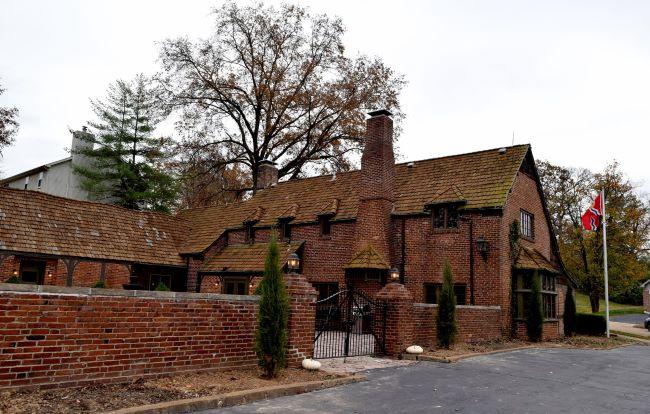 Historic brick house with a chimney, surrounded by a brick wall and small trees, under an overcast sky.