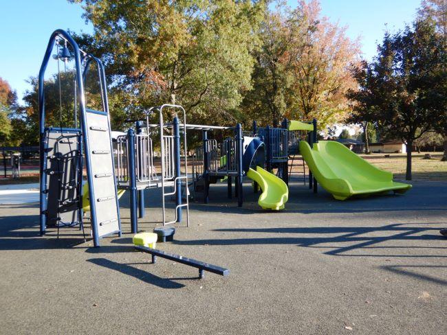 New playground with blue and green slides and climbing structures, surrounded by trees and autumn foliage.