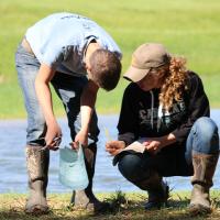 One student kneeling in front of a pond writing information on a piece of paper while another student bends over to observe.