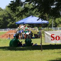 Students sit under a tree for shade while taking the soils test. A tent is located behind them. Beyond that a pit is dug in the earth with another student walking toward the pit.