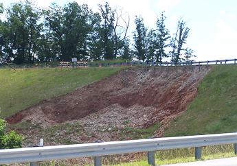 A section of surface soil and grass suddenly moved down a steep slope on the other side of a roadway guardrail.