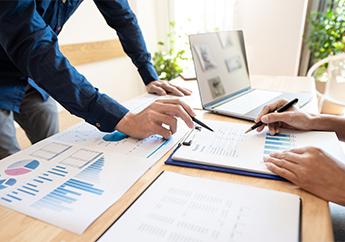 Two individuals working at a desk with a laptop and spreadsheets 