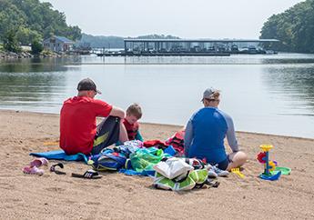 People sitting on a beach with beach gear surrounding them, looking across a lake with a large boat dock in the distance