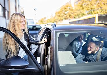 A woman entering a dark gray car through the open passenger car door while the male driver smiles and watches her.