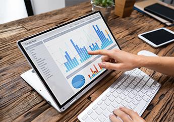 A person points at a graph on the screen of a laptop computer sitting on a wood desk..