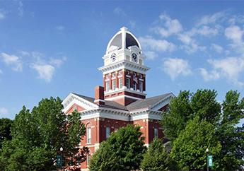 Top view of a two-story red brick building featuring a four-stage, square clock tower with a pyramidal slate roof atop the intersecting wings