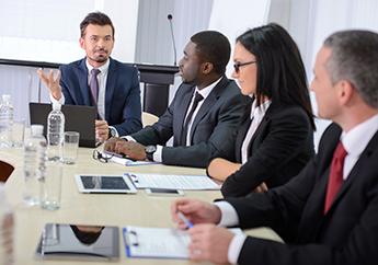 Several men and women wearing suits sitting at a large table with computers having a meeting