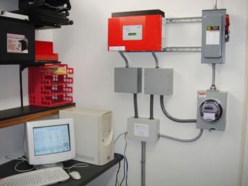 A personal computer sits on a desk. There are power boxes and an electric meter on the wall.