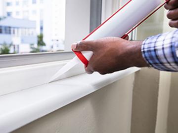 A man installs weatherization caulking around a window sill.