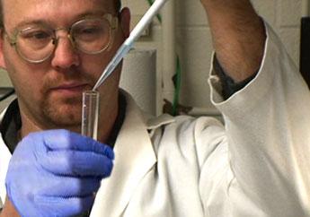 Laboratory worker wearing protective glasses putting liquid in a test tube vial using a dropper tool.