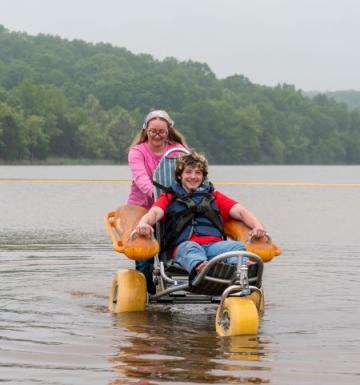 Smiling young man in adaptive beach chair enjoys being pushed through the water by a woman.