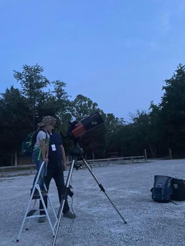 A child stands on a stepladder to look through a telescope as an adult assists.