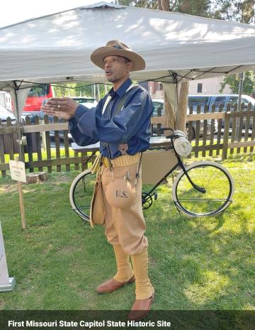 A Black man dressed in historical-period clothing standing in front of a bicycle from the 1800s.