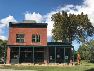 Two-story brick building with one-story addition to the side. Sign on one—story side says “BANK.”