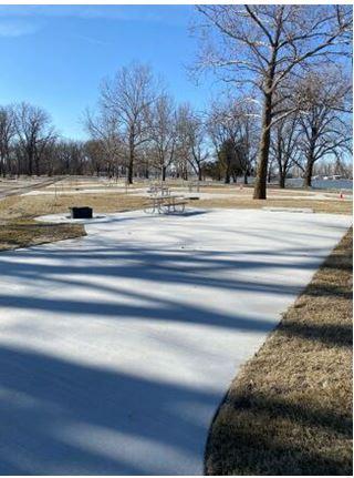 Concrete parking pads with picnic table and fire pit on them. Trees scattered throughout the camping area in front of a lake.