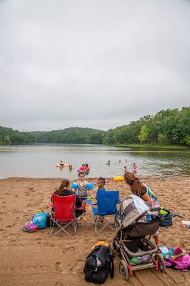 Beach on lake with swimmer. In foreground, woman standing near stroller and young children with floaties standing by chairs with female in camp chair