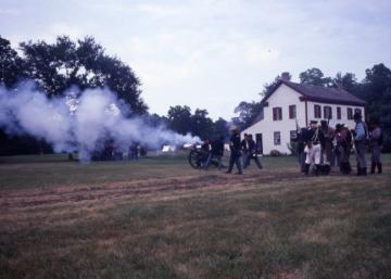 Civil War soldiers firing cannons and muskets outside a white house on a grassy field during a reenactment.