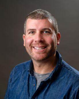 Fletcher Bone, a man with short hair wearing a denim shirt over a gray t-shirt, against a dark background.