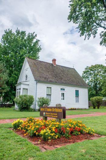 Small white siding home with Harry S Truman Birthplace on brown sign with yellow and red flower surrounding home and sign in front yard.