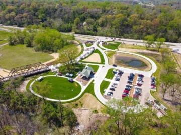 Aerial view of a park with winding paths, parking lot, pond, green lawn, trees, and a nearby road
