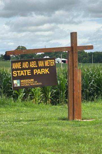Sign for Annie and Abel Van Meter State Park, mounted on a wooden post, set against a green cornfield and cloudy sky