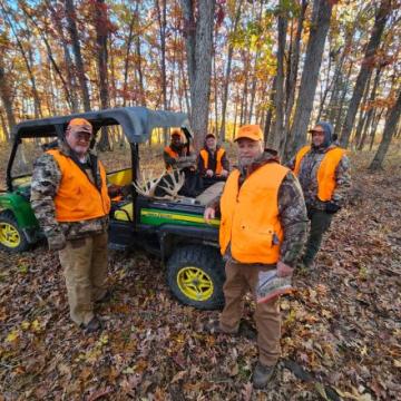 A group of five men in orange vests stands by a green utility vehicle in a wooded area during autumn, with a deer antler visible in the vehicle bed