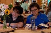 Two women seated at a table practice their nature journaling. Multicolor summer flowers in vases decorate the tables.