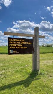 Brown cantilever sign that says Sappington African American Cemetery State Historic Site in  a grassy field under a blue cloud-filled sky.